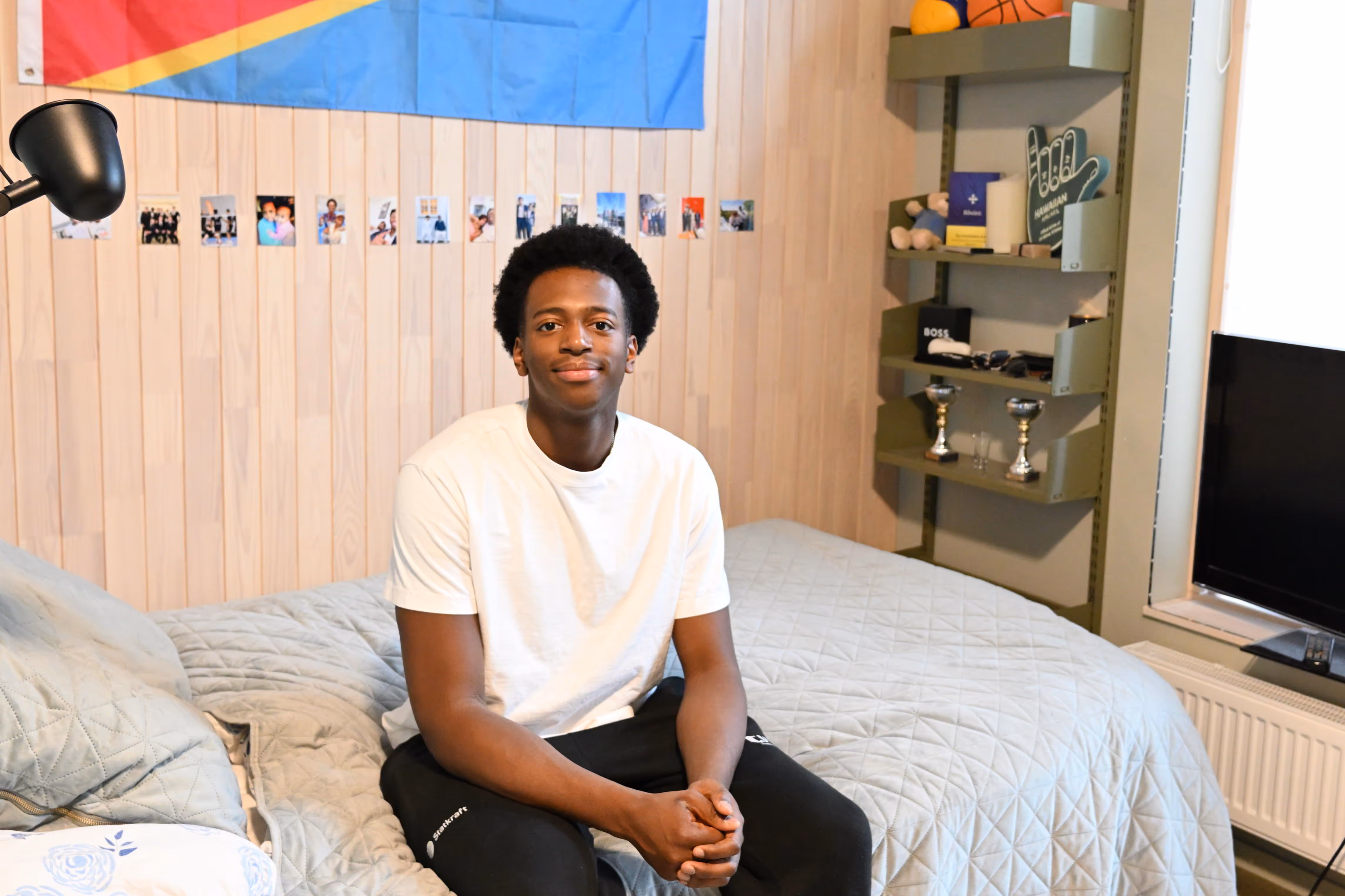 Ung mann i kvit t-skjorte som sit på ei seng i eit rom med lyse trepanelvegger, bilder på veggen og ei grønn hylle med trofé og ei TV.