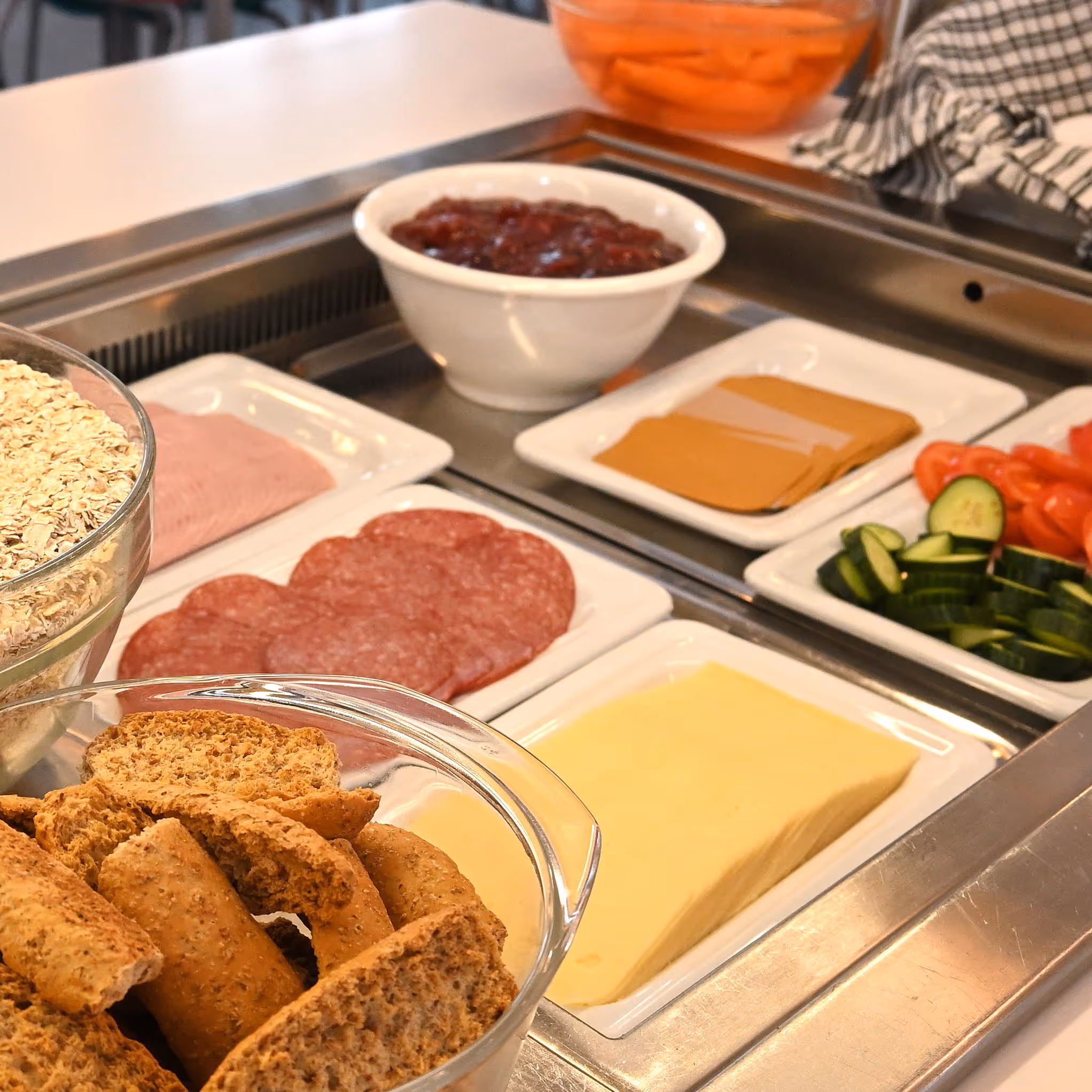 Frokoststasjon med knekkebrød, havregryn, skiver av ost, salami, kokt skinke, agurk, tomat og en skål med rød syltetøy.