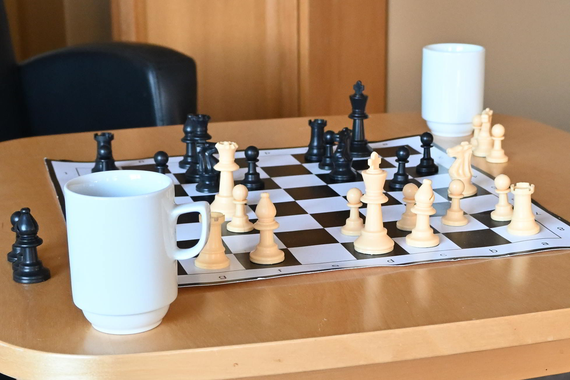 Et sjakkbrett med svarte og hvite brikker plassert på et bord, med to hvite kopper ved siden av brettet.