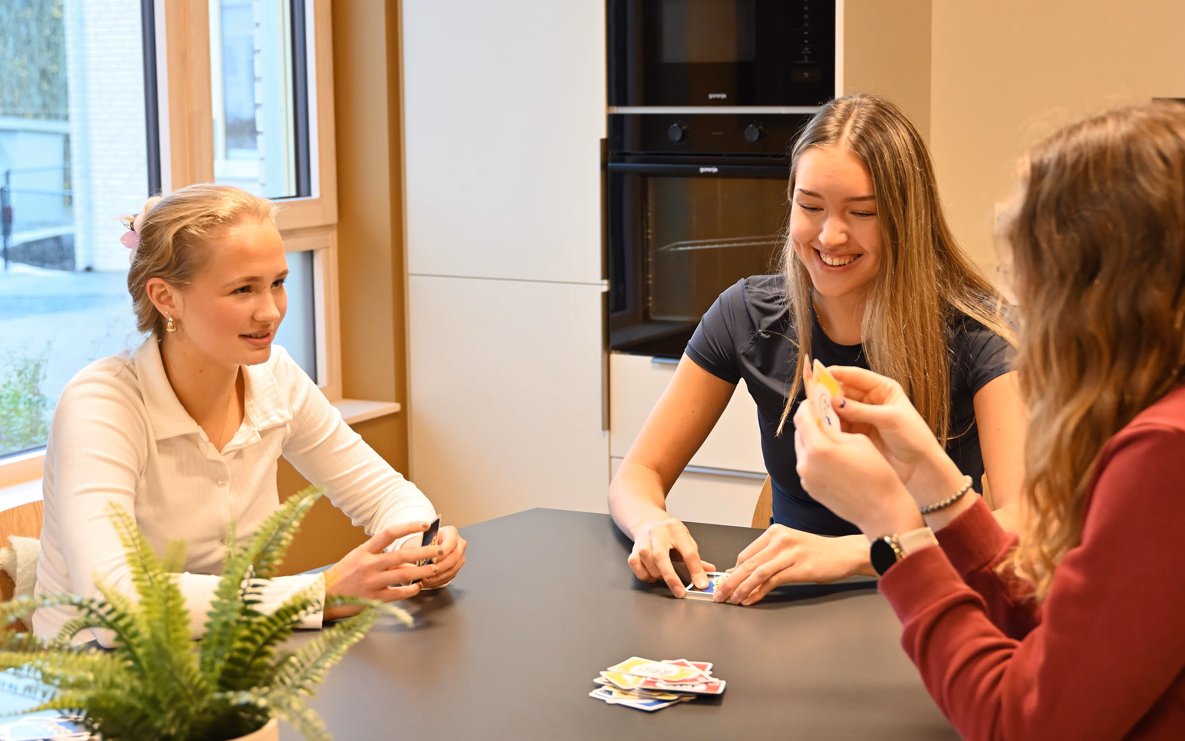 Tre ungdommer som sitter rundt et bord og spiller kortspill, ler og har det moro sammen.