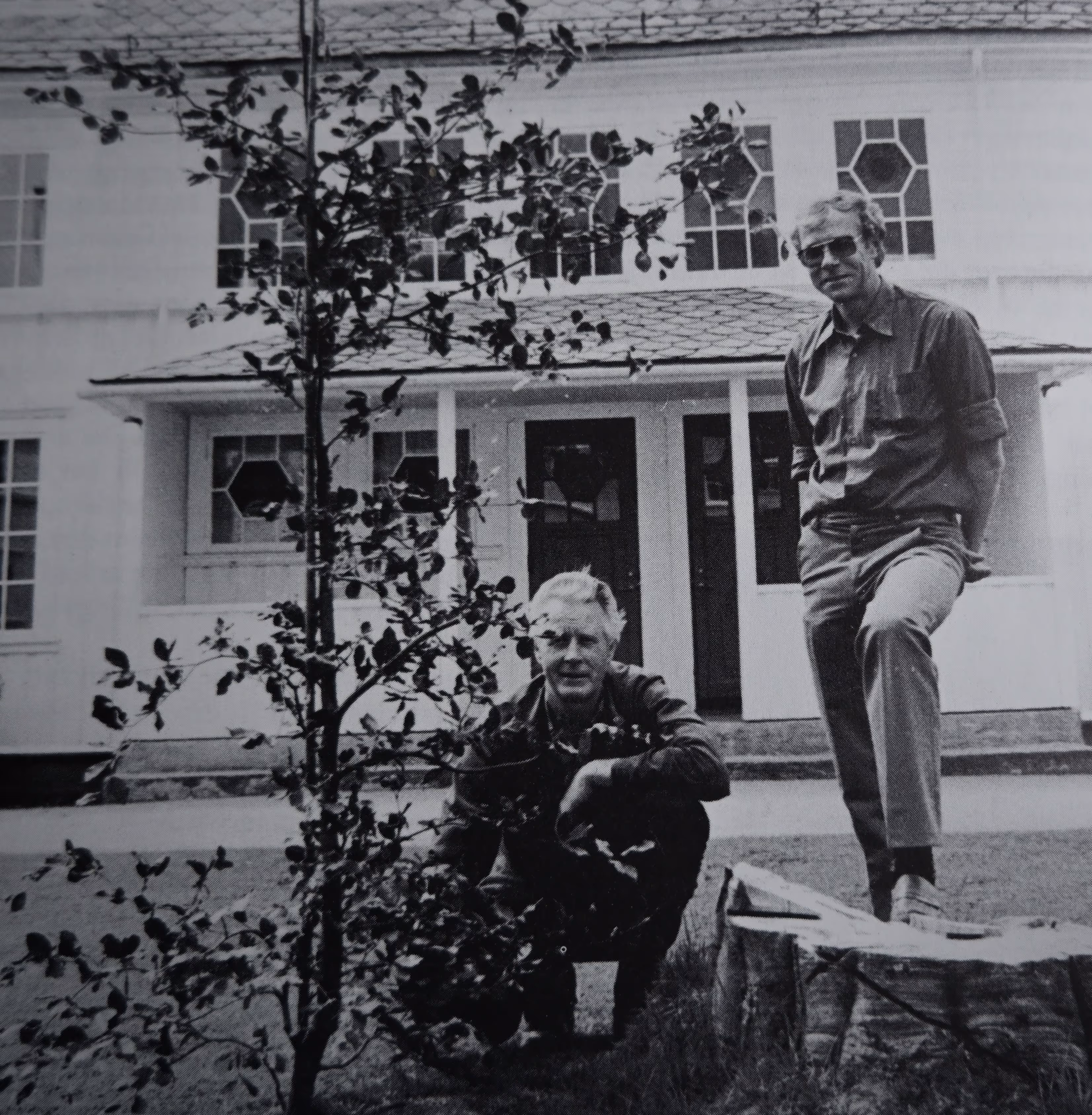 To menn med solbriller ved et lite tre foran et hus med dekorative vinduer.