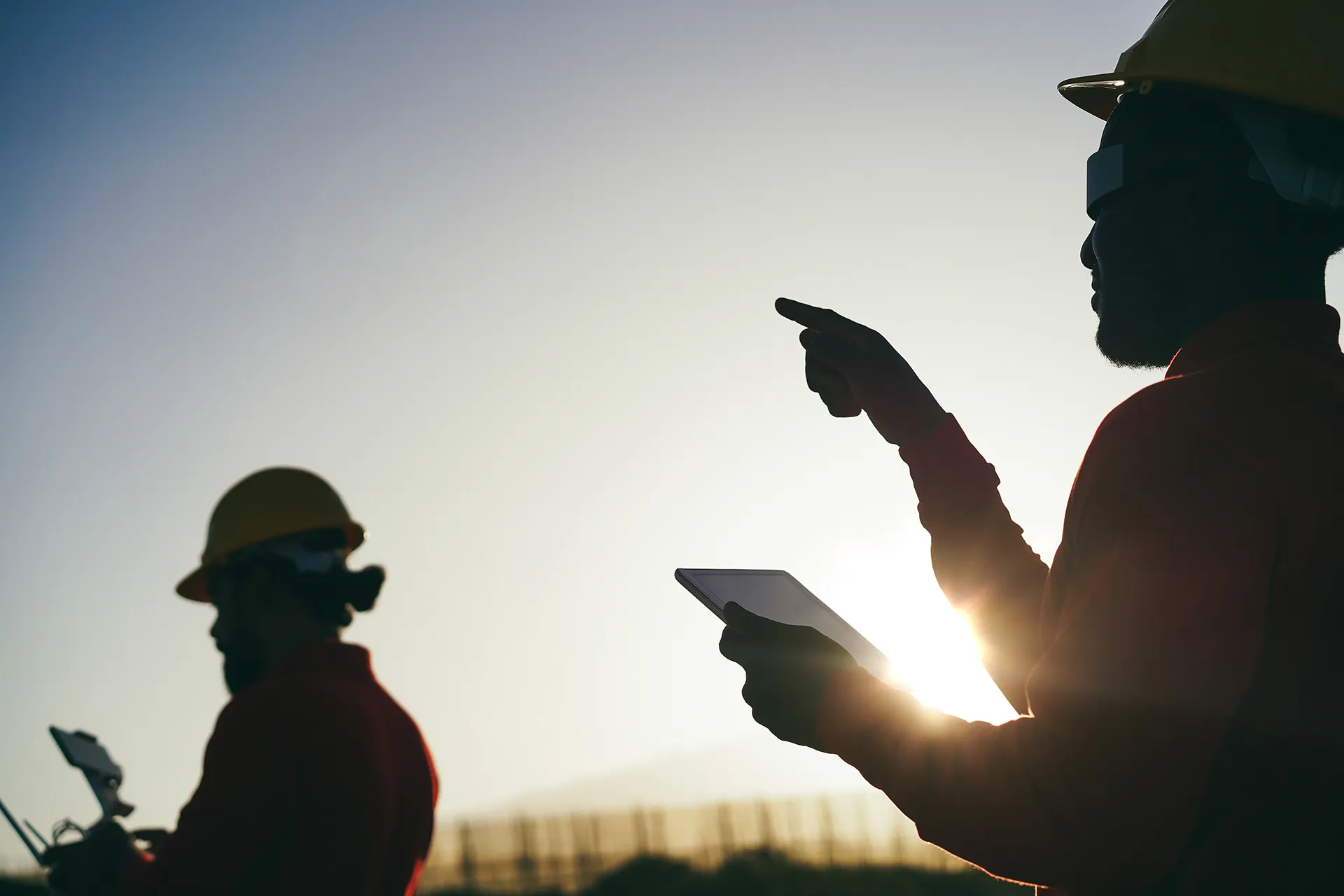 Two engineers wearing yellow hard hats using a tablet and a remote control outdoors during sunset.