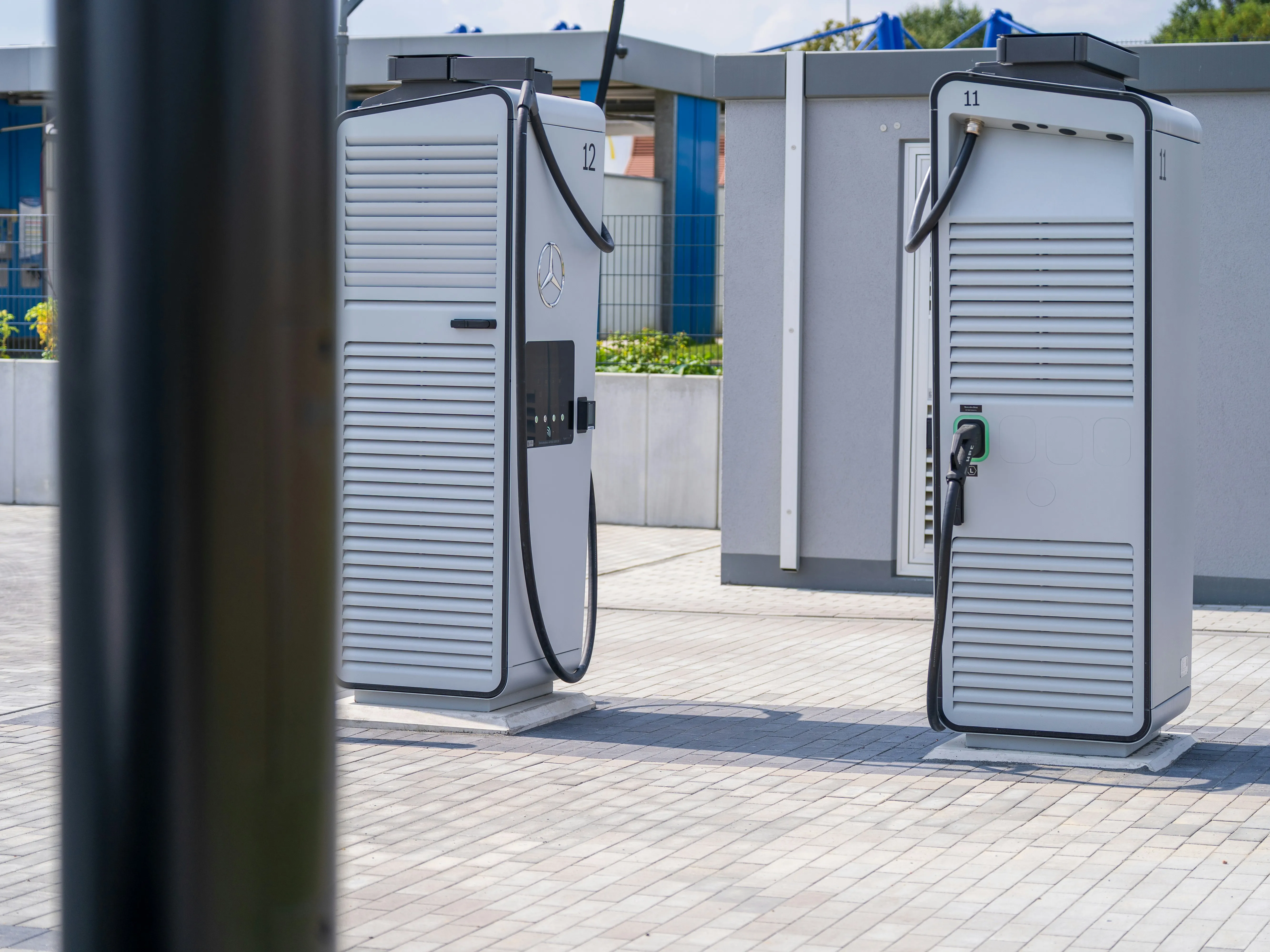 Two Mercedes electric vehicle charging stations on a tiled pavement near a gray building.