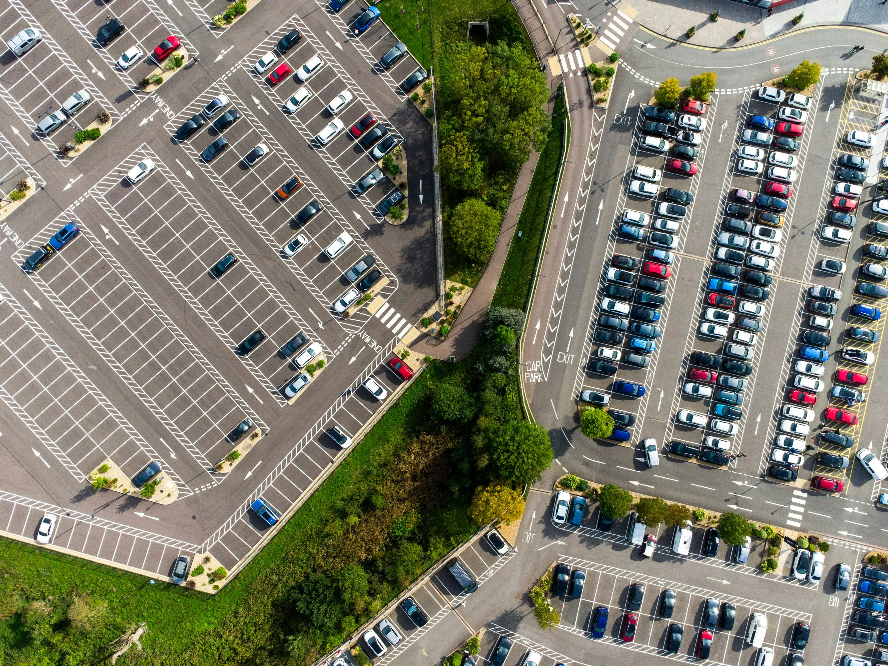 Aerial view of a large parking lot with many parked cars and a small green space with trees in the middle.