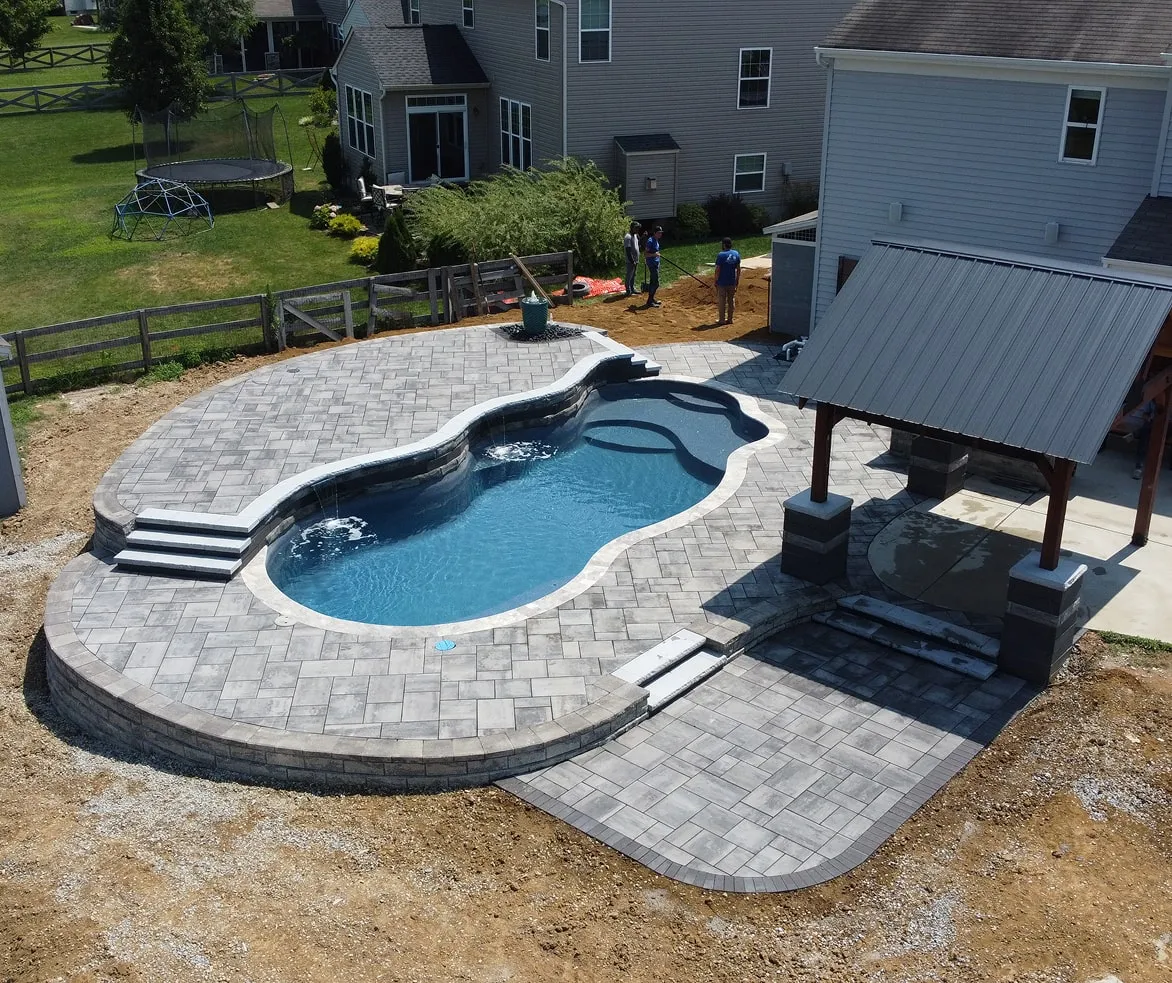 Backyard with a kidney-shaped pool surrounded by grey stone pavers, a covered patio area, and workers preparing the ground near a two-story house.