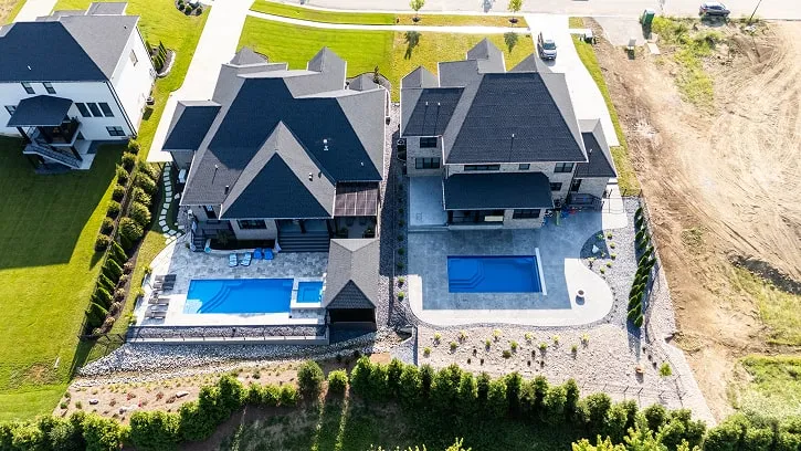 Aerial view of two modern houses with dark roofs, each featuring a blue swimming pool in spacious backyards with stone landscaping.