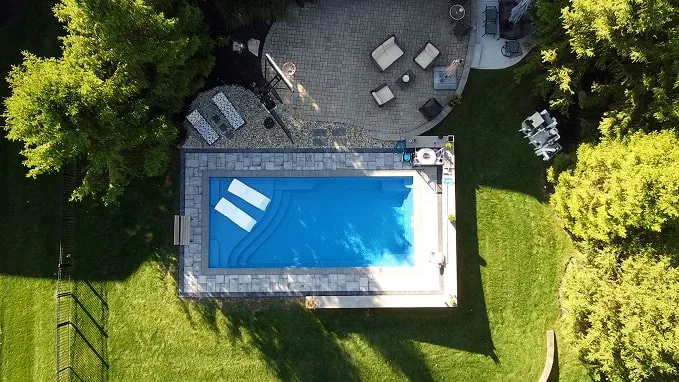 Aerial view of a rectangular swimming pool with two white lounge floats in a backyard surrounded by green grass and trees.