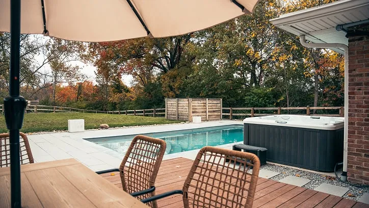 Backyard scene with a rectangular pool, hot tub, wooden deck with chairs, and large umbrella, surrounded by autumn trees.