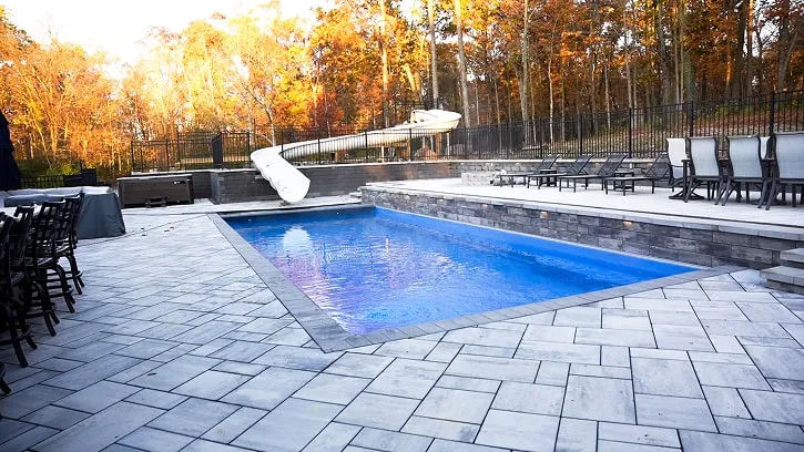 A backyard swimming pool with a white slide, surrounded by lounge chairs, dining chairs, and autumn trees in the background.