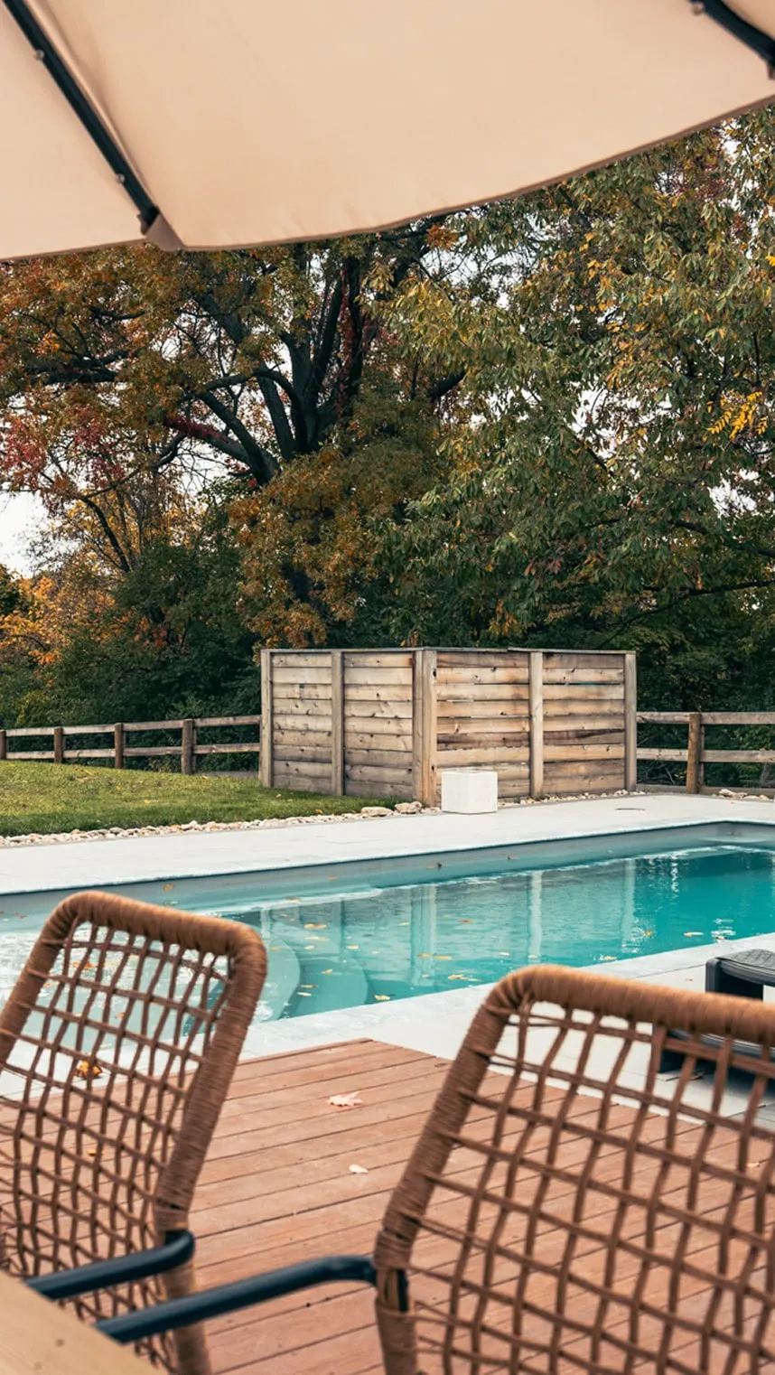 Outdoor pool with clear blue water, wooden deck with woven chairs, and autumn trees in the background.