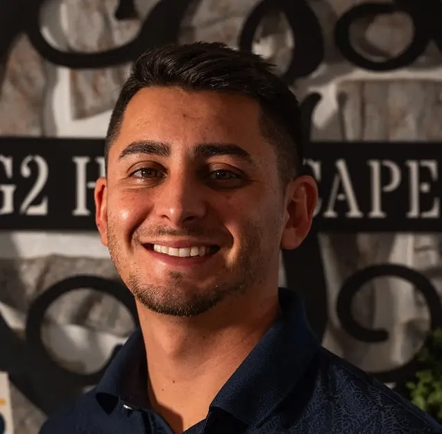 Smiling man with short dark hair and trimmed beard wearing a dark collared shirt in front of a stone wall with partially visible black and white sign.