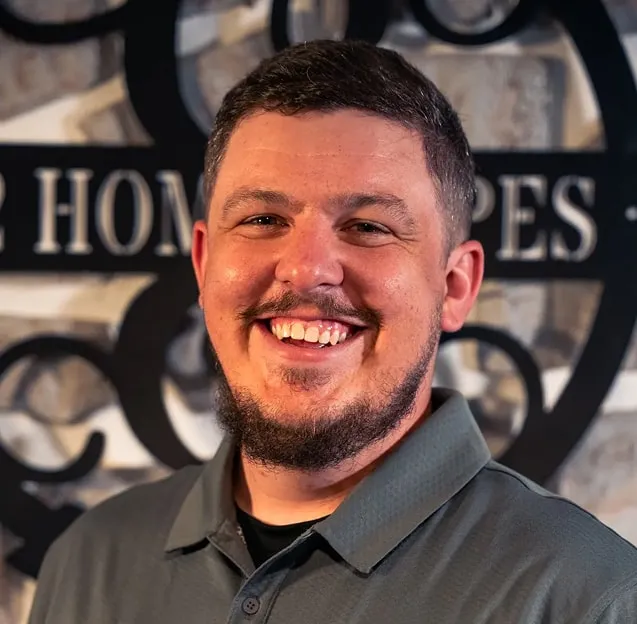Smiling man with short dark hair and a beard wearing a gray collared shirt, standing in front of a decorative sign.