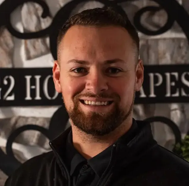 Smiling man with short hair and beard wearing a dark jacket standing in front of a stone wall and decorative metal sign.