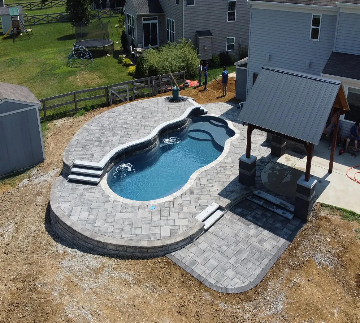 Backyard swimming pool with gray stone paver deck, small waterfalls, and a covered patio area next to a house.