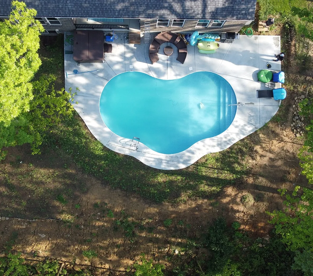 Aerial view of a kidney-shaped swimming pool with surrounding concrete patio, outdoor seating, and greenery.