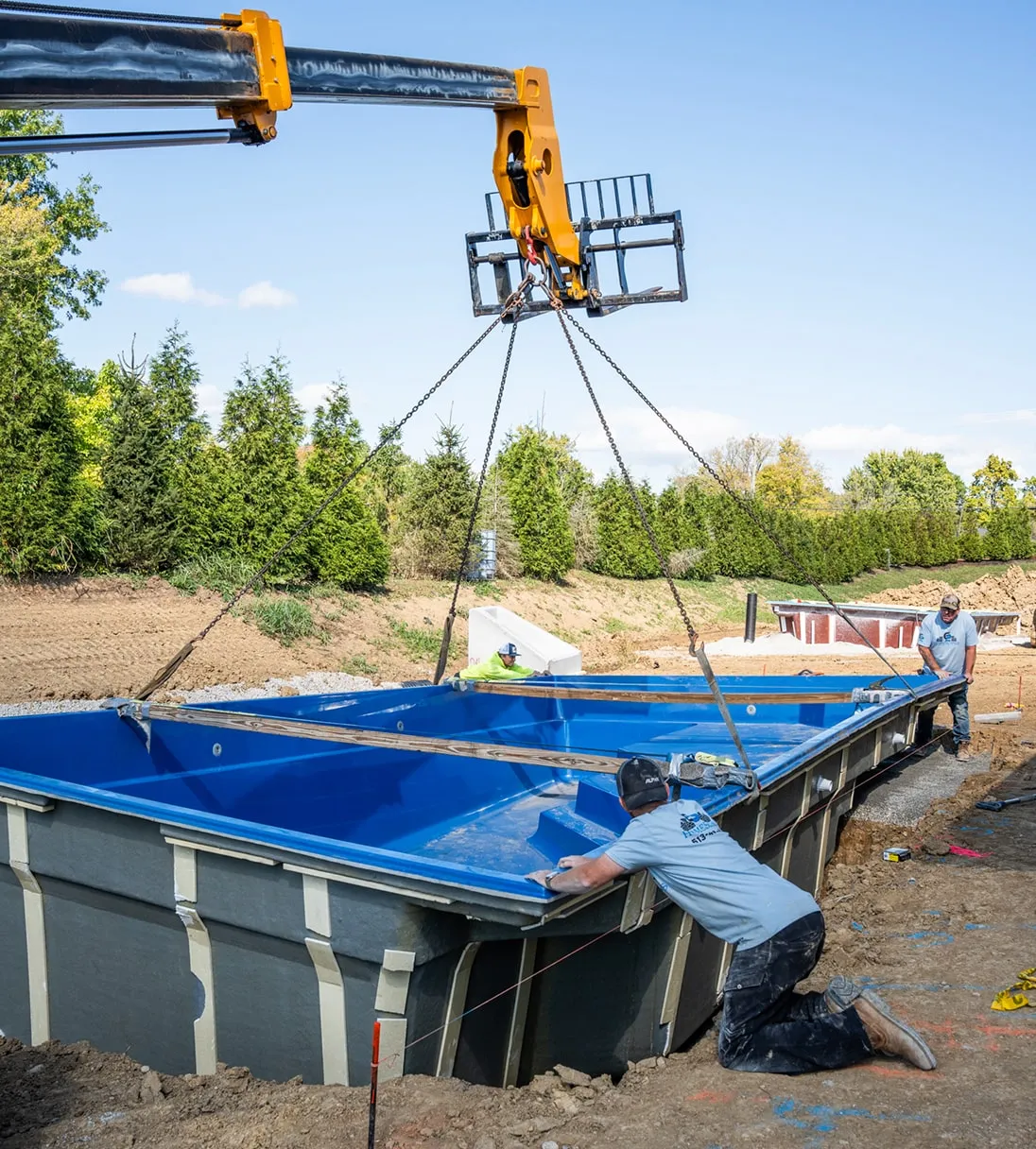 Construction workers installing a large blue prefabricated pool shell into a dug hole using a crane on a sunny day.