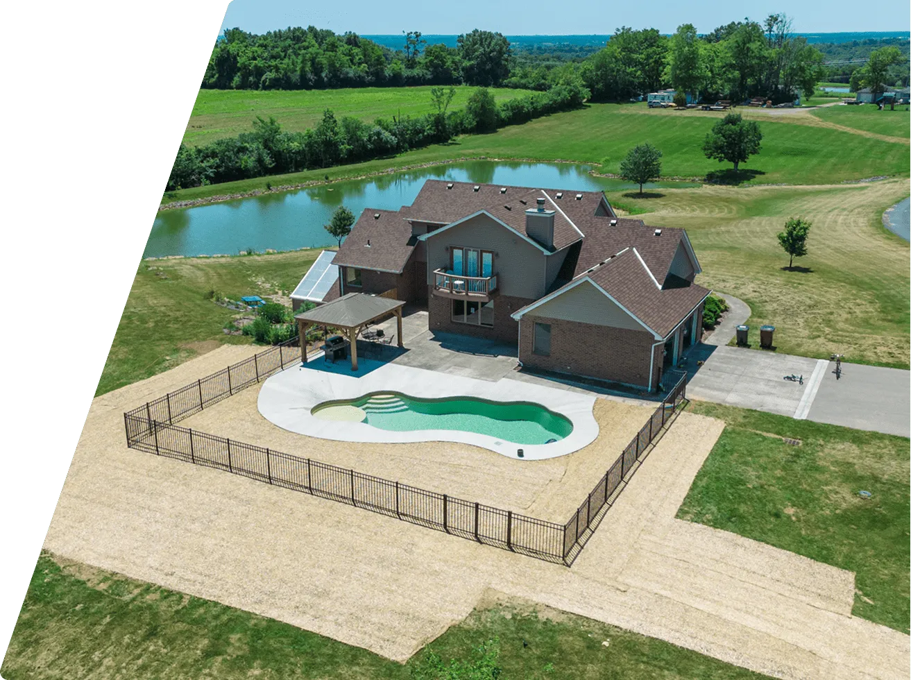 A large brick house with a fenced kidney-shaped swimming pool in the backyard, surrounded by a gravel area and green fields with a pond nearby.