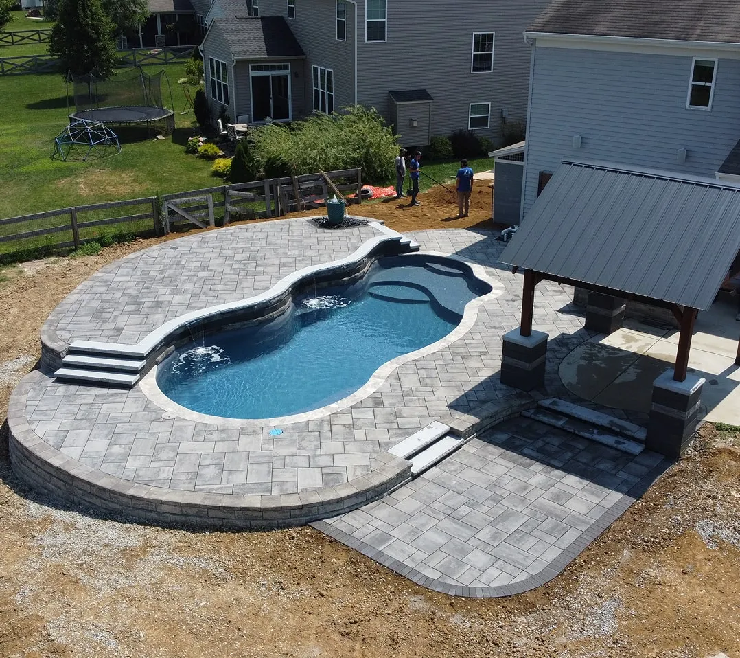 Backyard with a kidney-shaped swimming pool surrounded by gray stone pavers, a covered pavilion, and three people working near a house.
