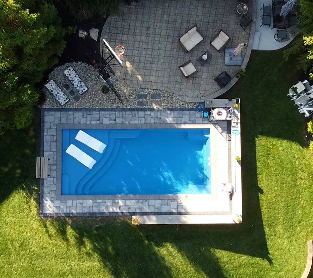 Aerial view of a rectangular backyard swimming pool surrounded by stone tiles with two white lounge chairs in the water, adjacent patio area with outdoor seating, and green lawn.