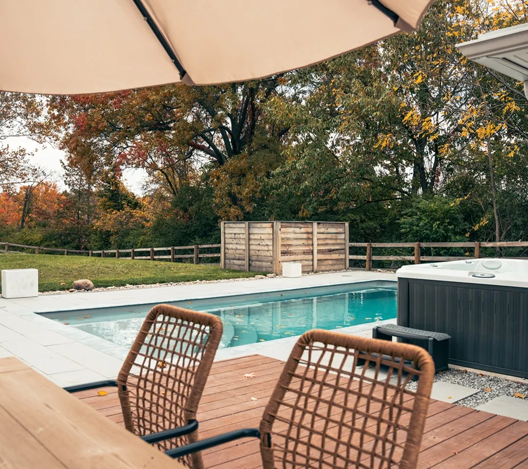 Backyard with a rectangular pool, wooden deck with two wicker chairs, a hot tub, and autumn trees in the background.