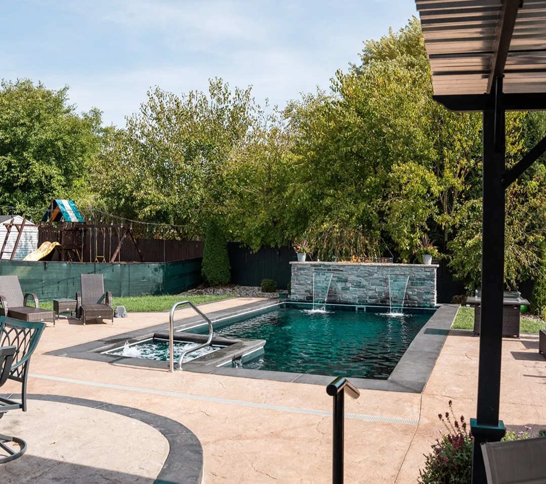 Modern backyard with a rectangular swimming pool, water fountains, spa area, patio furniture, and green trees in the background.