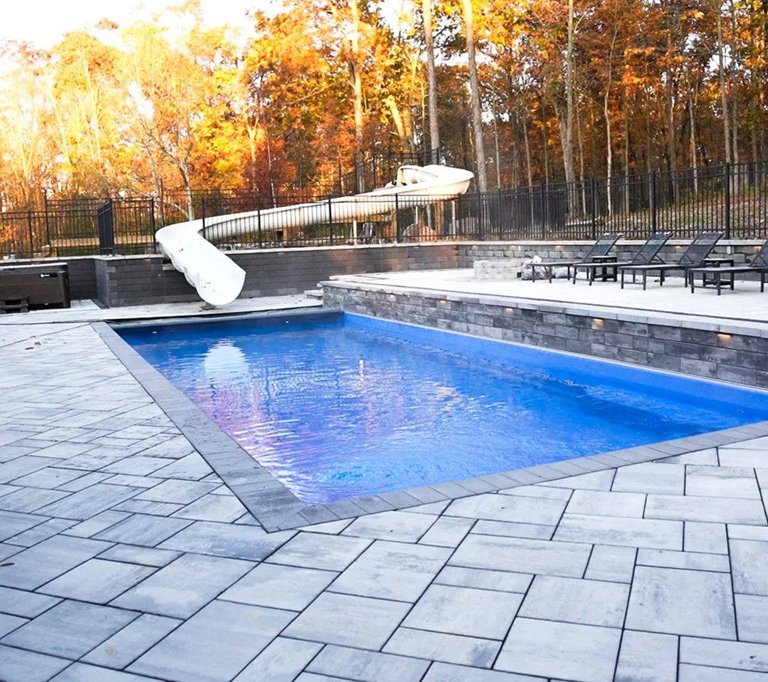 Outdoor swimming pool with a white spiral water slide, surrounded by gray stone patio and several lounge chairs, set against autumn trees.
