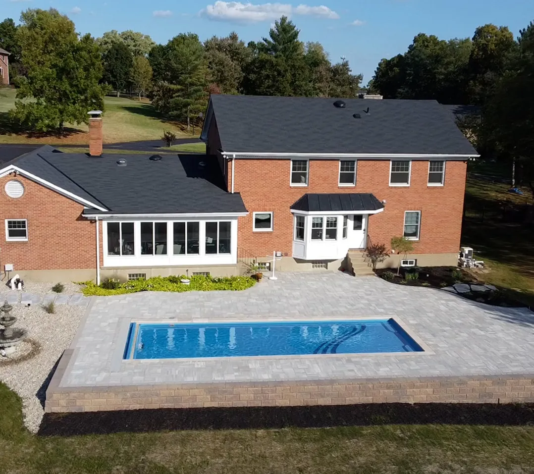 Brick house with a black roof and a rectangular in-ground swimming pool surrounded by light gray stone patio.