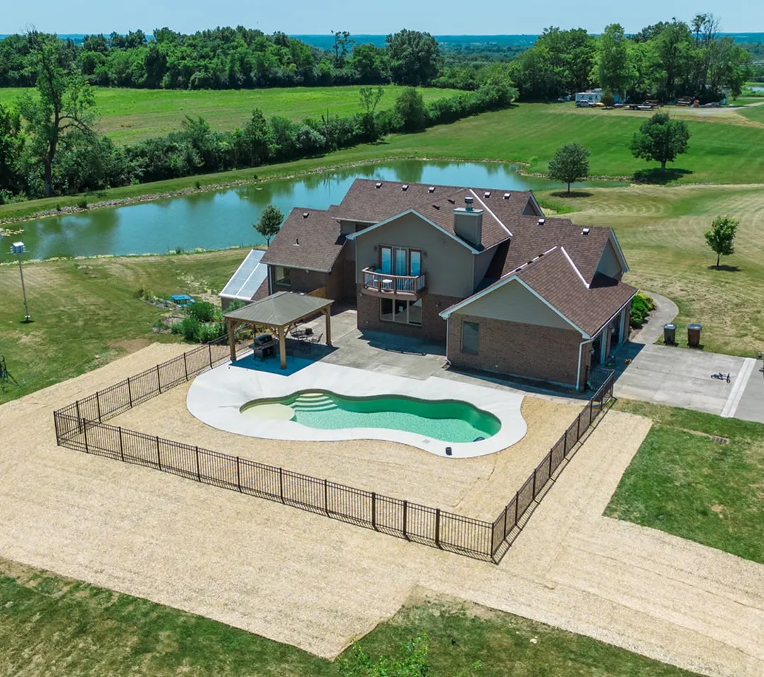 Aerial view of a large brick house with a fenced kidney-shaped swimming pool and a small gazebo, set in a spacious green lawn with a pond in the background.