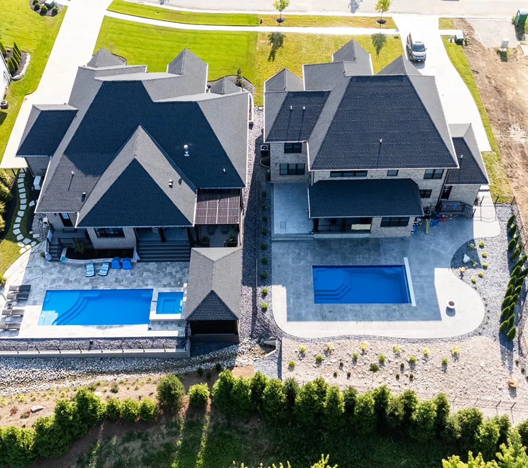 Aerial view of two modern houses with dark roofs, each featuring a rectangular swimming pool in their backyards.