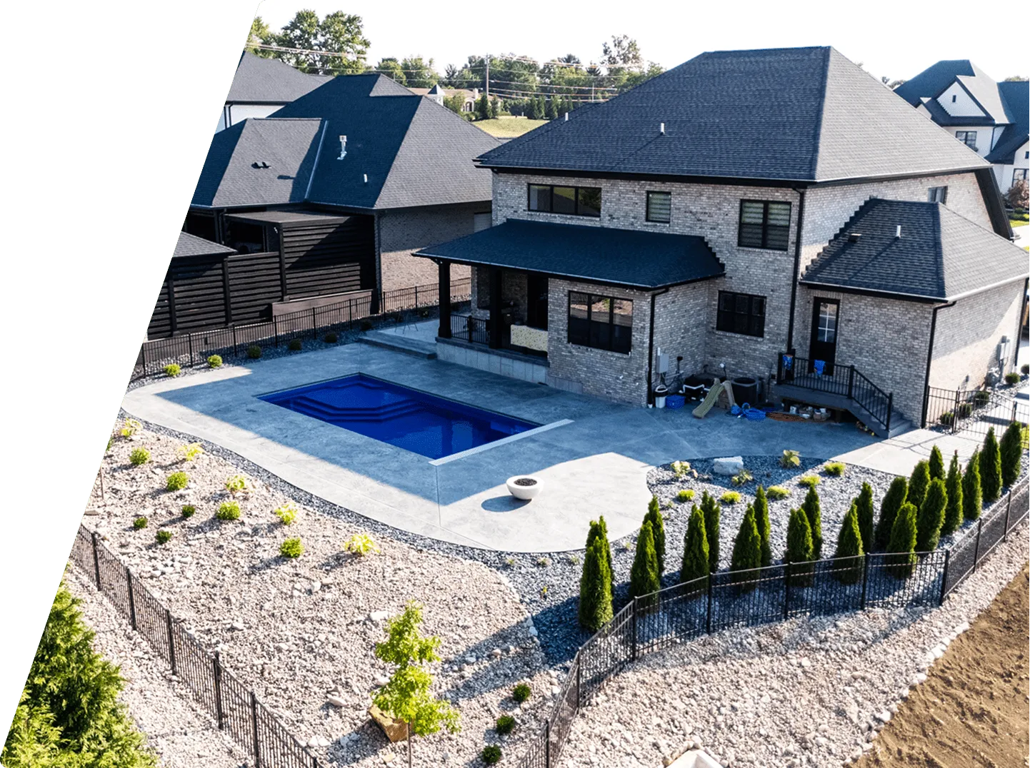 A modern two-story brick house with a dark roof, adjacent to a small rectangular blue swimming pool and landscaped yard with stone mulch and neatly trimmed shrubs.