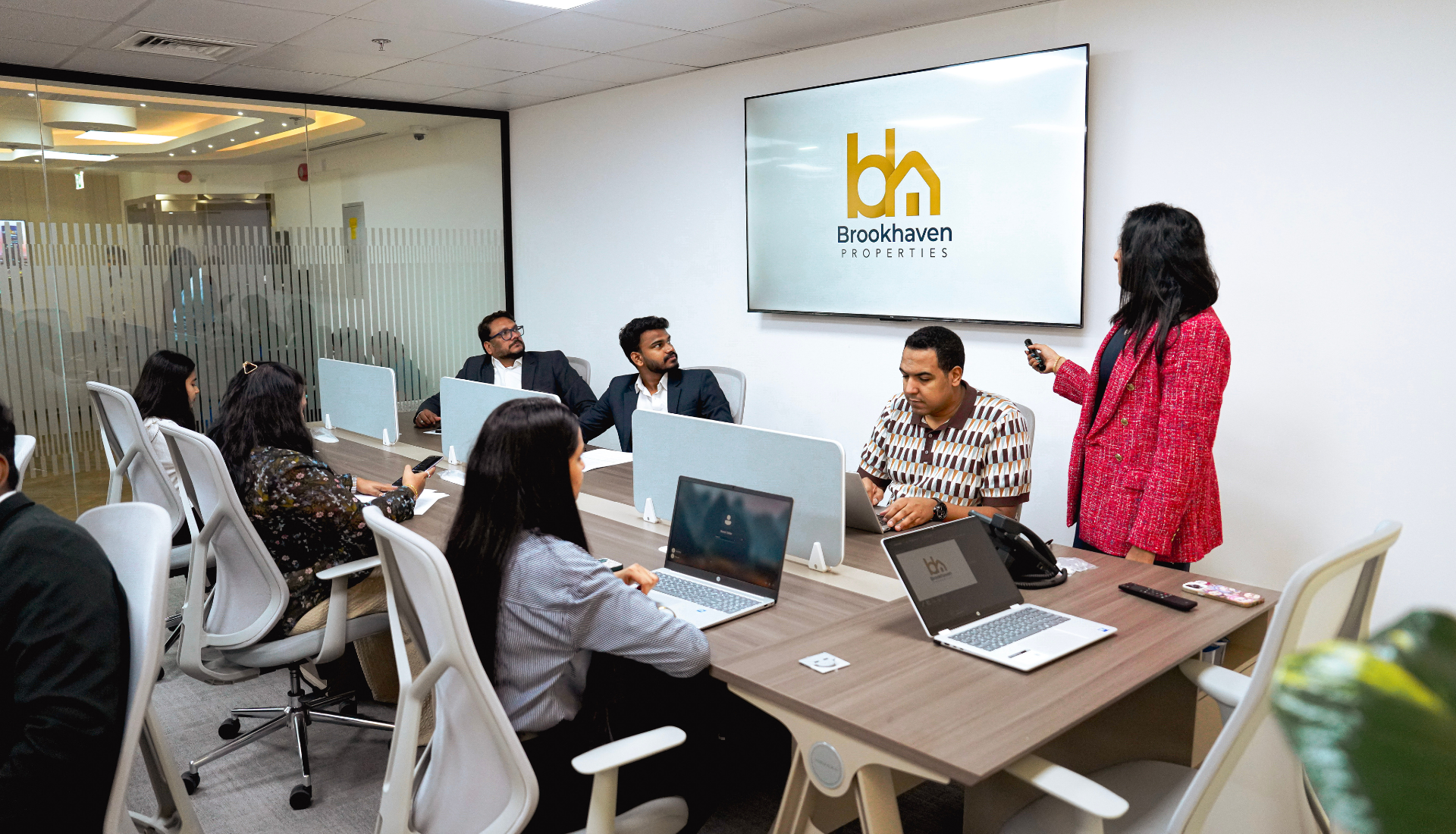 A business meeting in a modern conference room with seven people seated around a table, a woman in a red jacket is presenting using a large screen displaying the Brookhaven Properties logo.