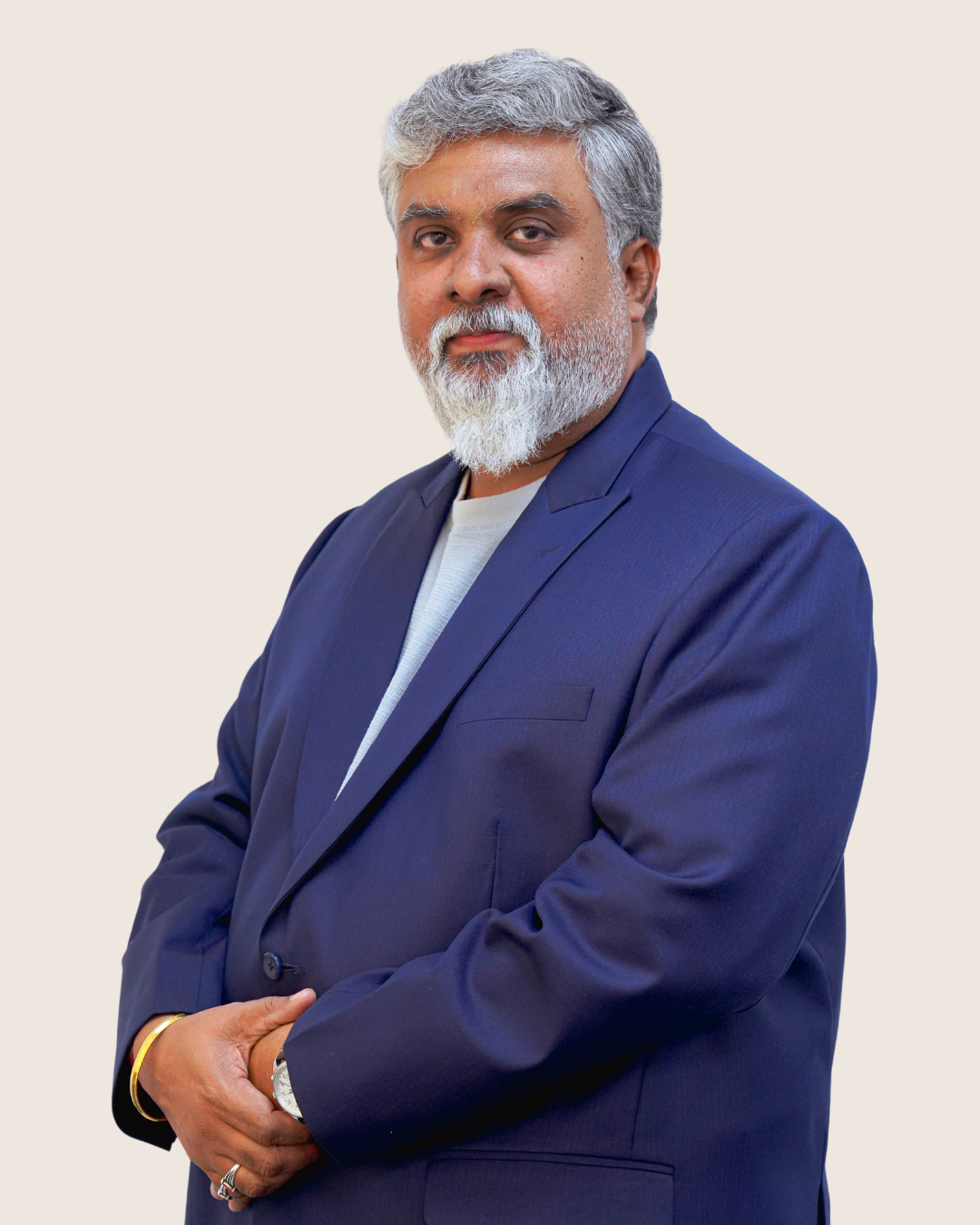 Middle-aged man with gray hair and beard wearing a navy blue suit jacket and white shirt, standing with hands clasped.