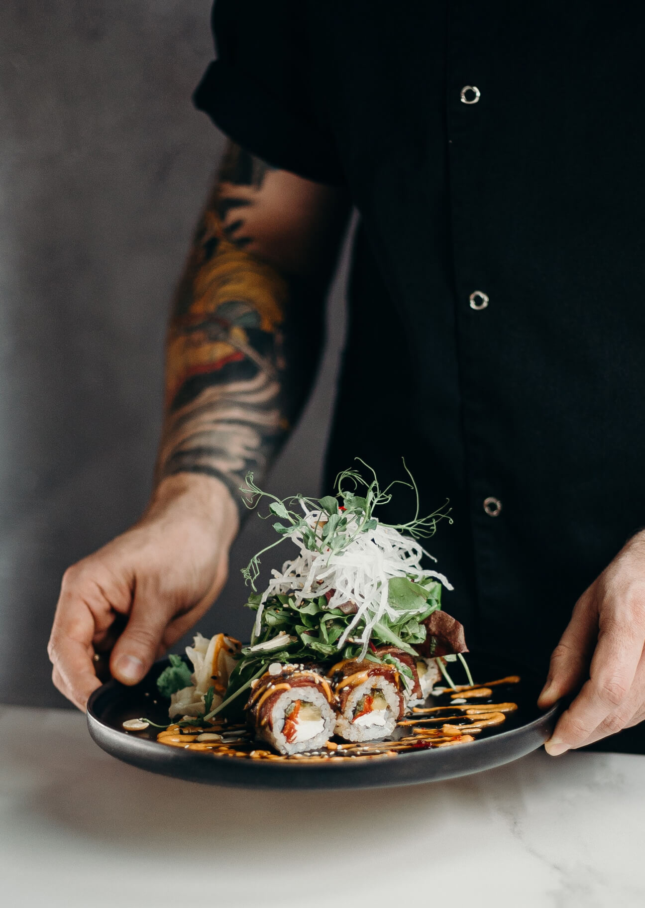Persona con tatuaje en el brazo sosteniendo un plato con rollos de sushi decorados con salsas, hojas verdes y tiras de rábano blanco.