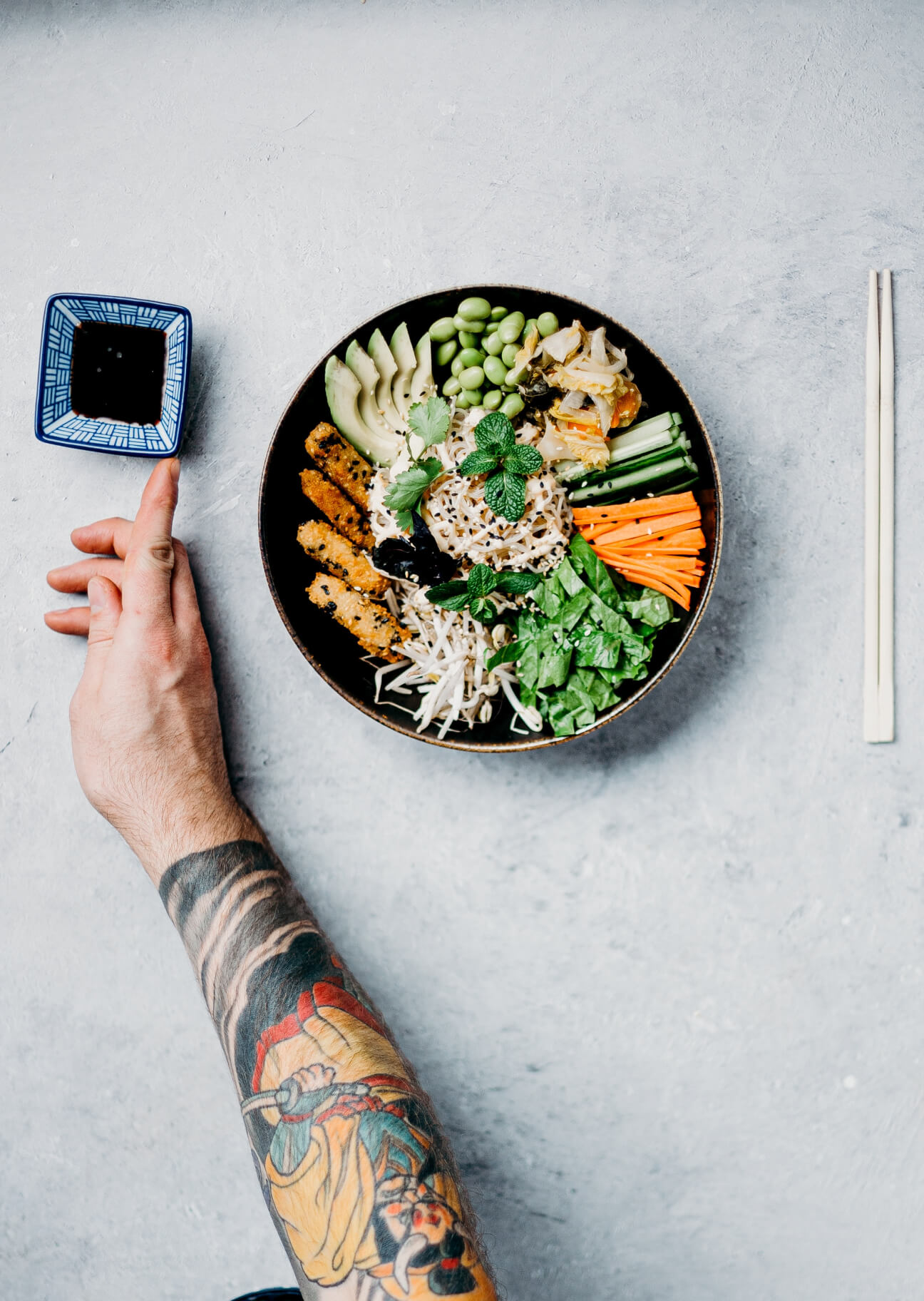Brazo tatuado sosteniendo un pequeño cuenco cuadrado con salsa junto a un bol de verduras, fideos y tempura con palillos a la derecha.