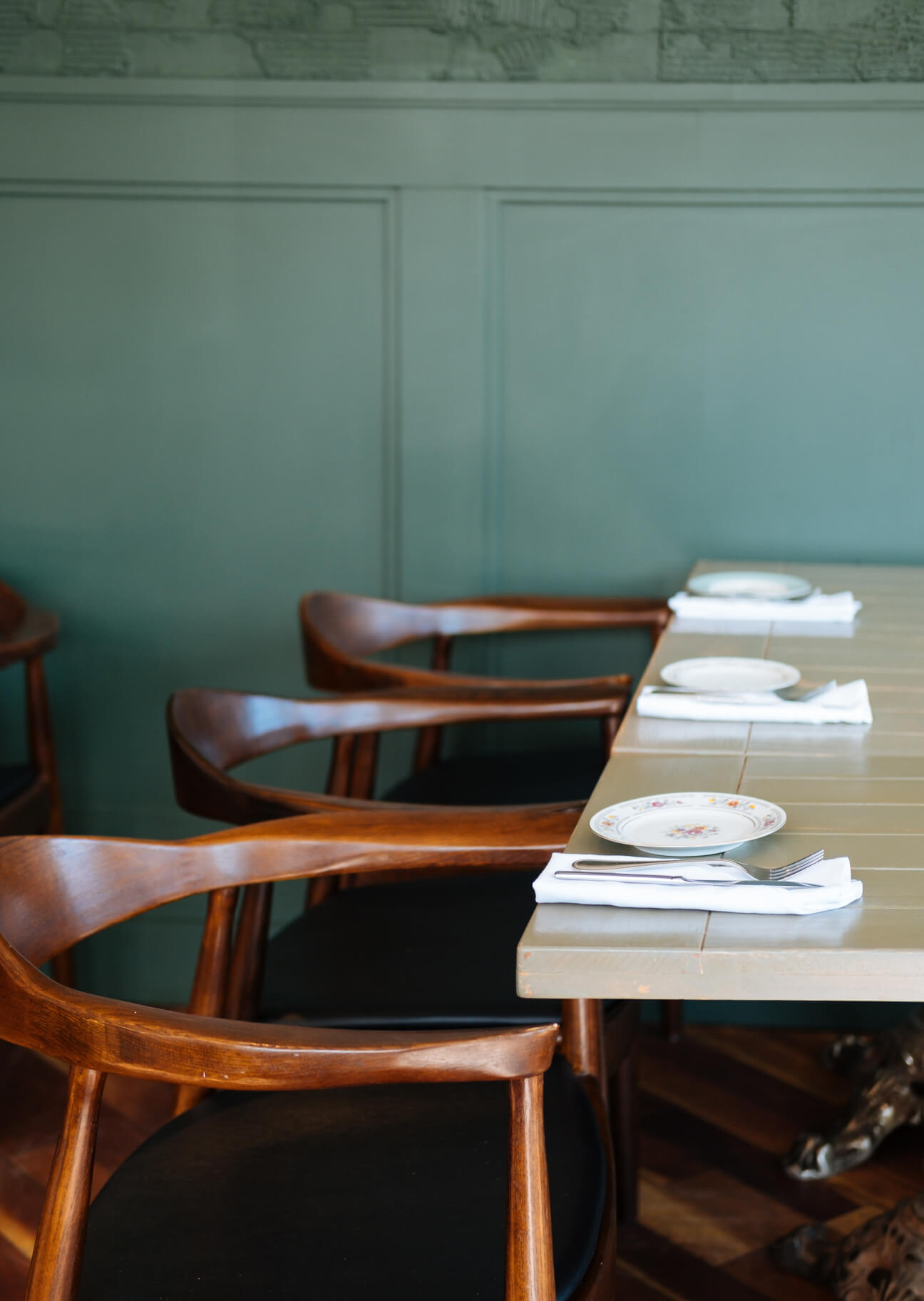 Mesa de restaurante con platos decorativos, servilletas blancas y cubiertos, junto a sillas de madera con asiento negro.