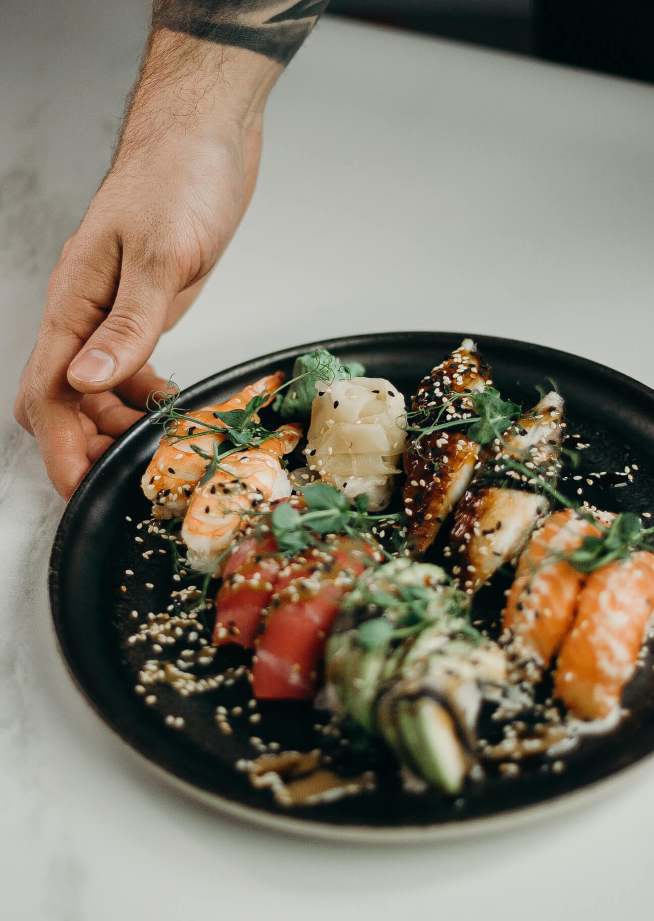 Plato negro con variedad de sushi decorado con semillas de sésamo y brotes verdes, con una mano masculina alcanzando el plato.