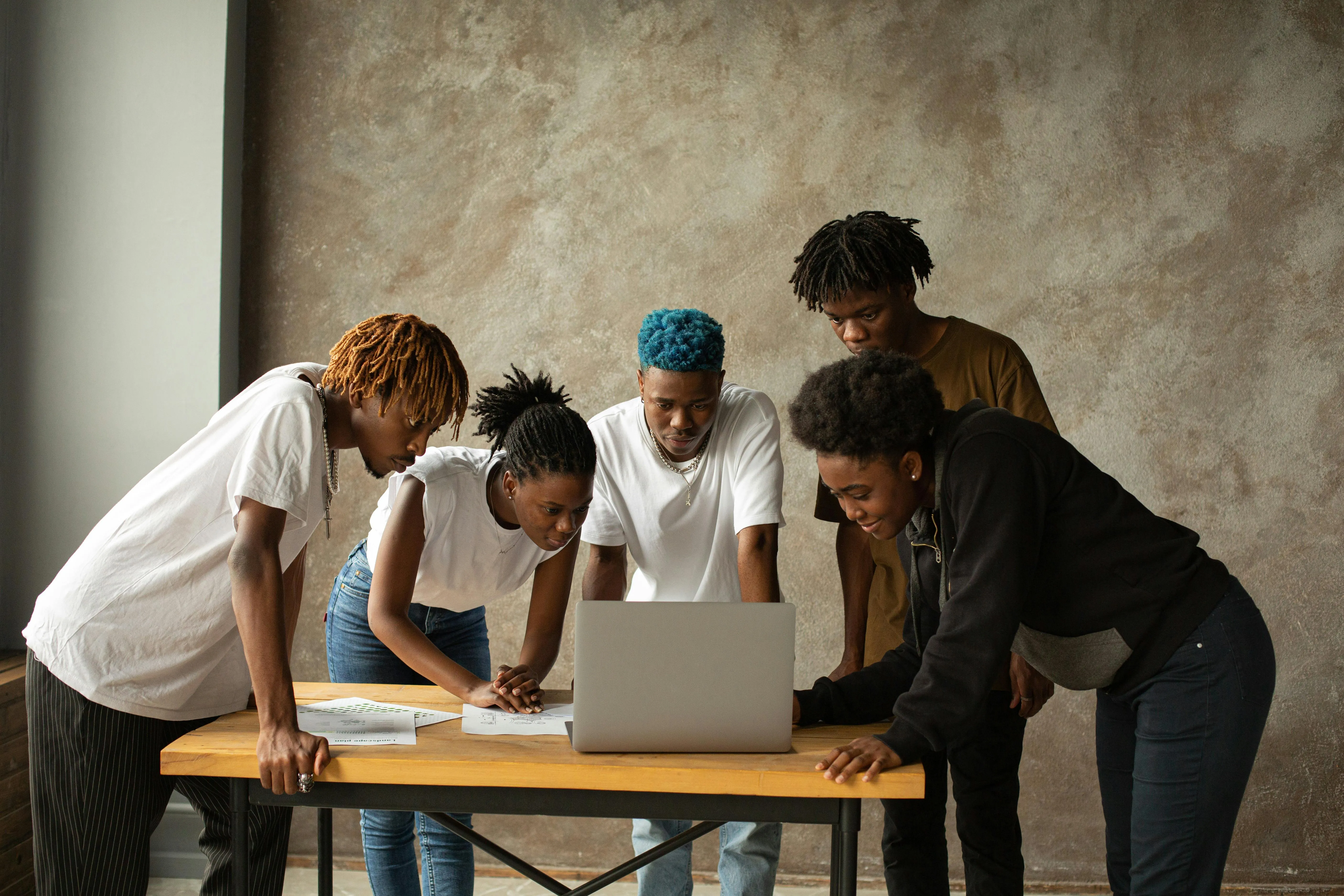 Five young people gathered around a wooden table looking at a laptop and papers.