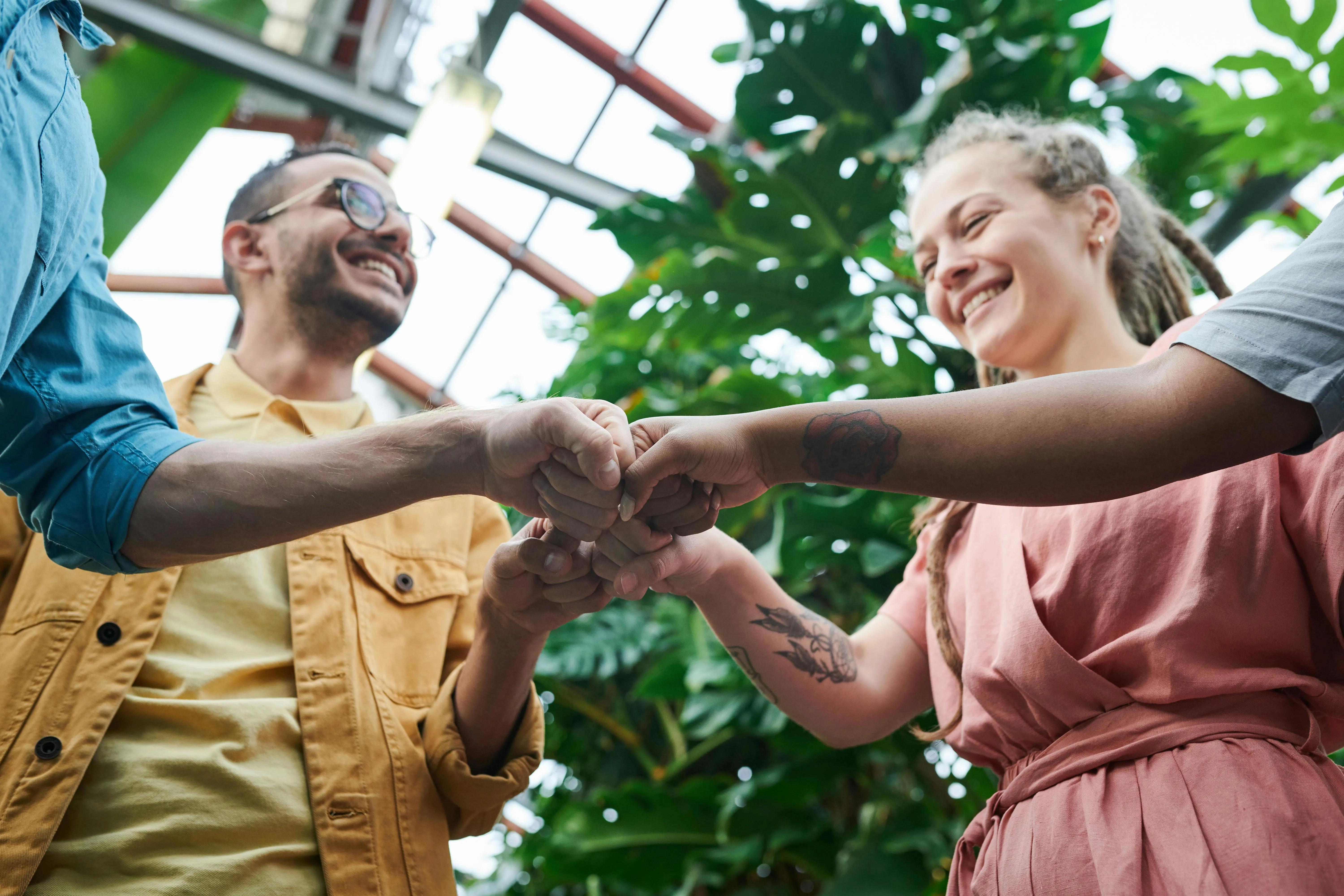 Smiling diverse group of people in casual clothes fist bumping in a bright indoor garden setting.