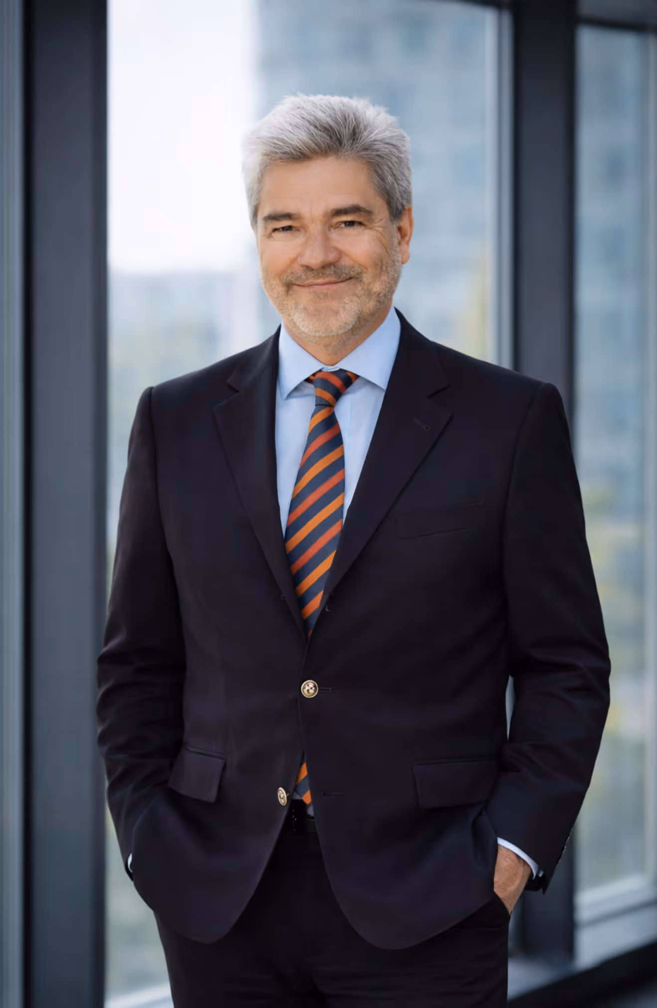Älterer Mann mit grauen Haaren in dunklem Anzug und gestreifter Krawatte steht vor einem Fenster und lächelt.