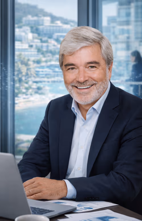 Älterer Mann mit grauem Haar und Bart lächelt am Schreibtisch vor Laptop in Büro mit Fensterblick auf Stadt und Wasser.