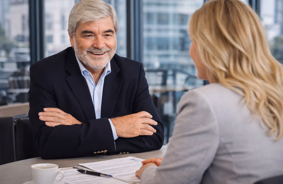 Älterer Mann im Anzug, der bei einem Gespräch lächelnd die Arme verschränkt, gegenüber einer blondhaarigen Frau in einem Büro.