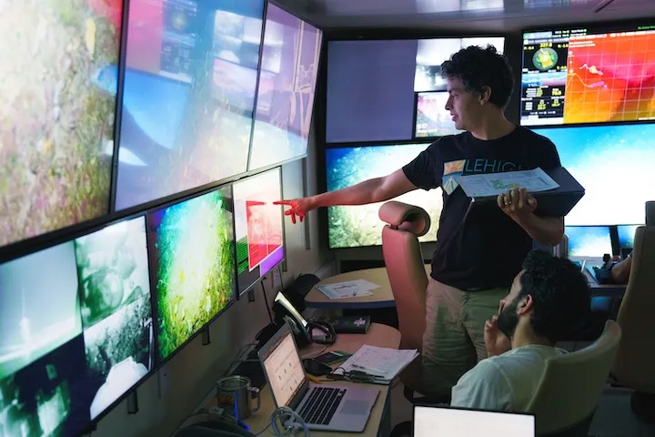 Two men in a control room with multiple large screens displaying underwater and data visuals, one pointing at a screen while the other looks on.