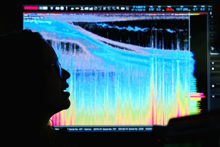 Silhouette of a person wearing glasses in front of a colorful, detailed sonar or scientific data display screen.