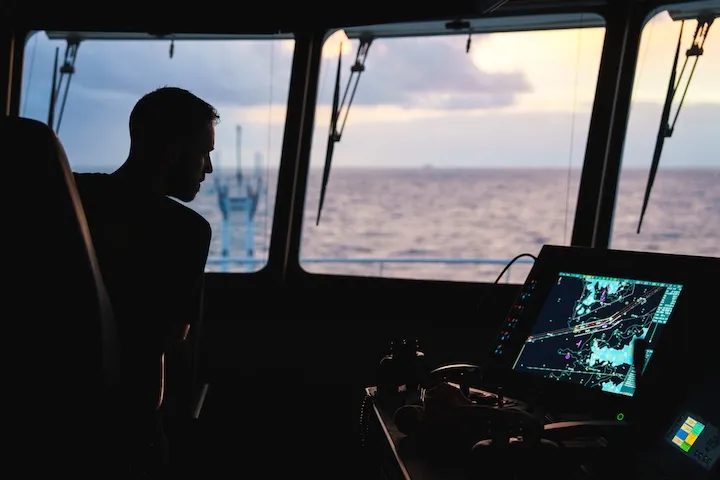 Silhouette of a person inside a ship's bridge looking out over the ocean during sunset, with a navigation screen displaying a map.