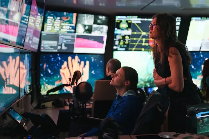 Two women monitoring multiple large screens displaying underwater coral and data in a control room.