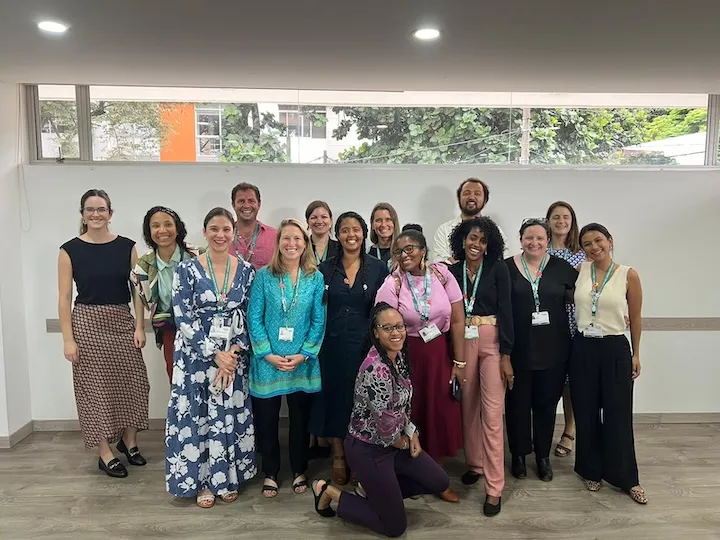 Group of sixteen diverse people standing and kneeling indoors in front of a window and white wall, smiling at the camera.