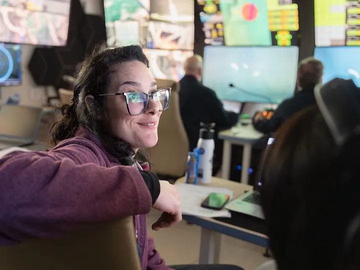 Woman with glasses smiling and sitting in a control room with computer monitors and colleagues in the background.