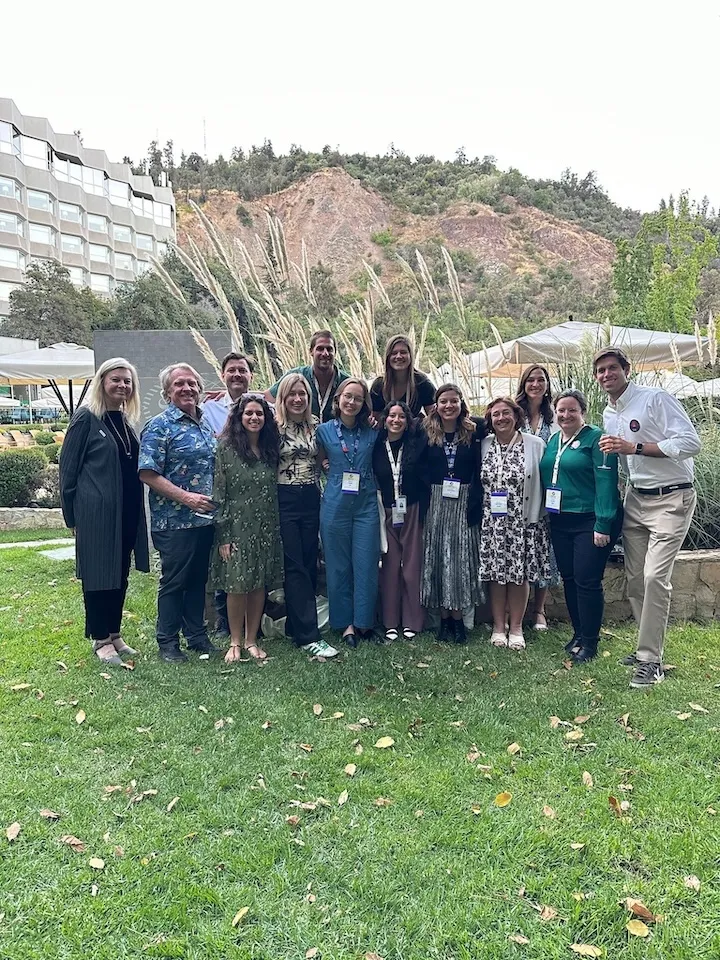 Group of fifteen people posing on green grass outdoors with hills and a modern building in the background.