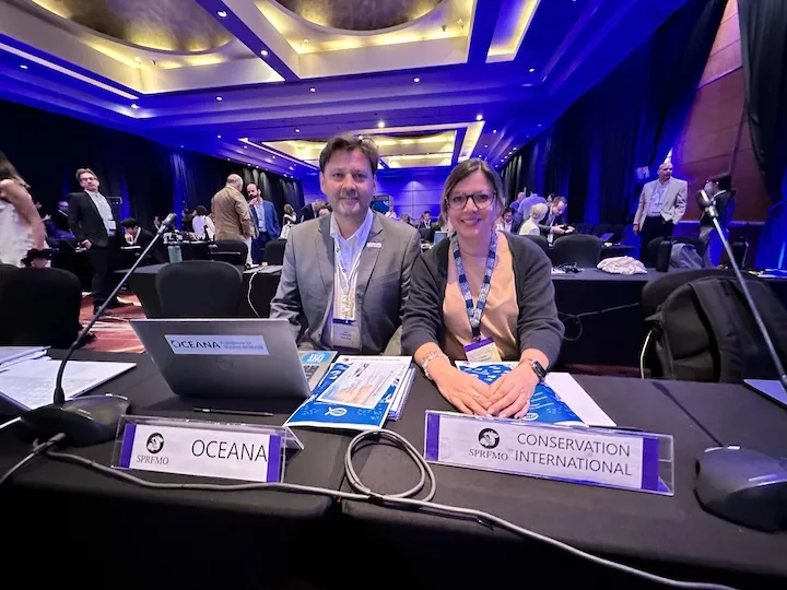 Two people seated at a conference table with nameplates labeled Oceana and Conservation International, surrounded by a large room of attendees.