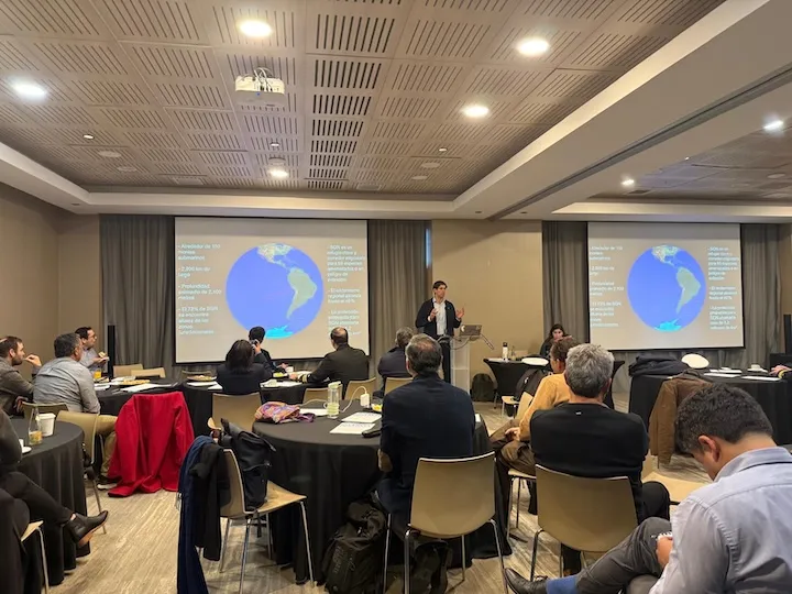 A speaker presenting data about global warming to an audience seated at round tables in a conference room with two screens showing a map of the Americas and climate-related statistics.